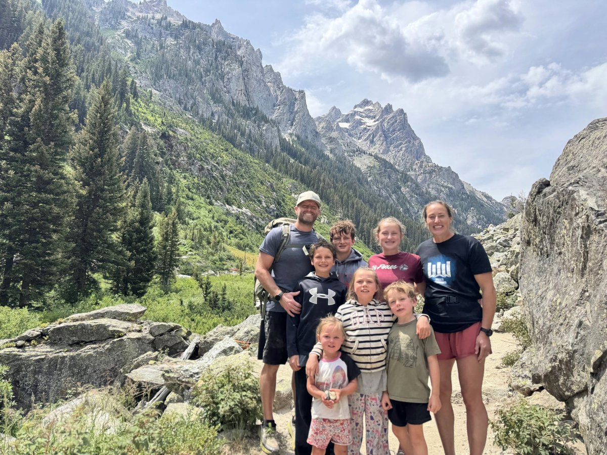 Family group of nine posing together on a mountain trail with forested peaks and rocky terrain in the background
